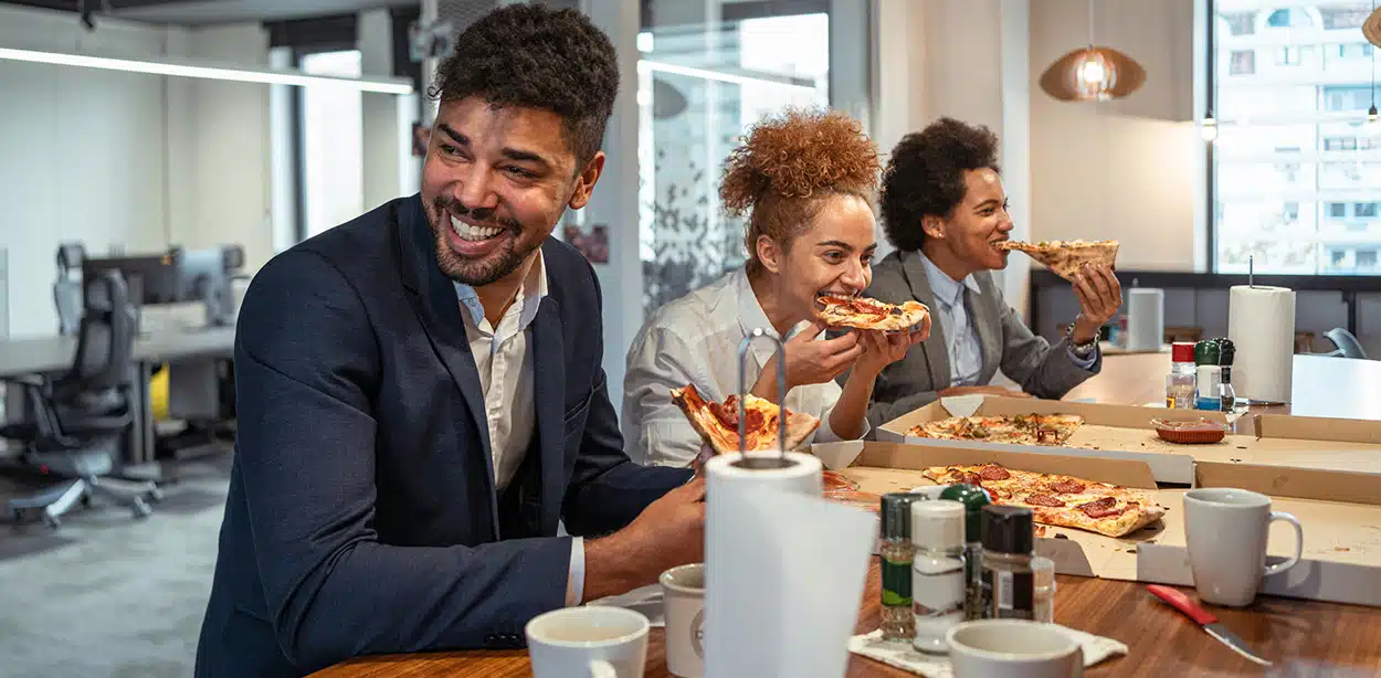Três pessoas rindo e comendo pizza no intervalo do escritório discutem sobre seguros.