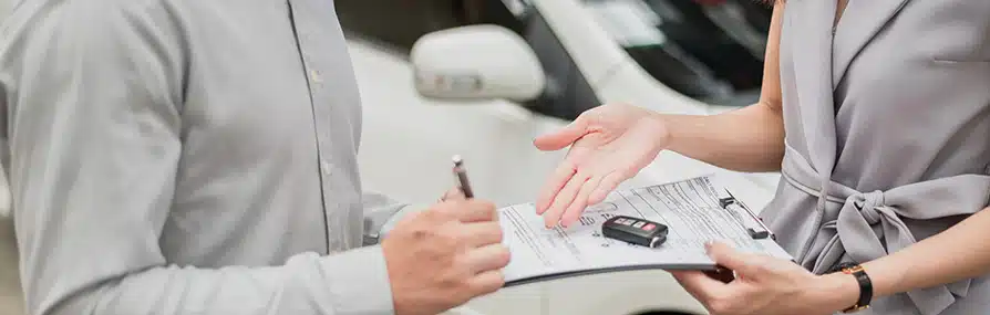 Duas pessoas próximas uma assina documento sobre seguros com chave de carro visível.