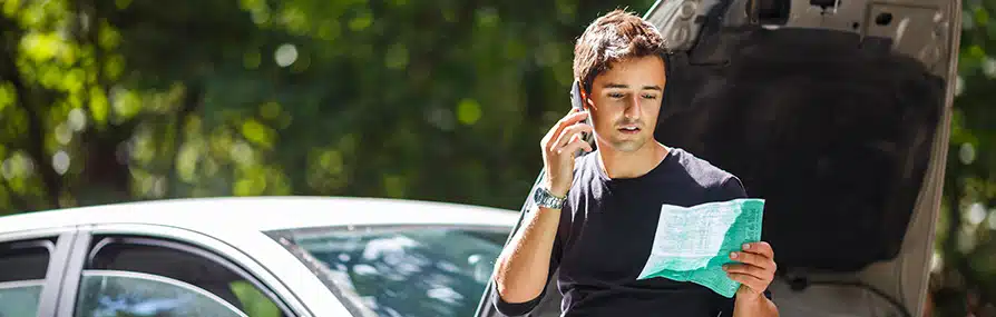 Jovem ao telefone com papel discute sobre seguros com capô do carro aberto ao sol.