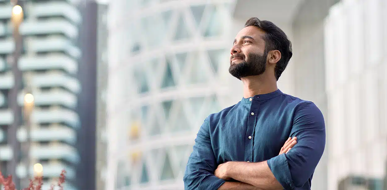 Homem sorridente de camisa azul e barba pensa sobre seguros com fundo urbano desfocado.